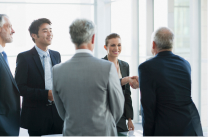 A group of five professionals in formal attire engage in a positive business meeting. A woman smiles while shaking hands, conveying a collaborative atmosphere.