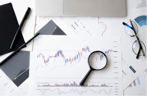 A desk with financial charts and graphs scattered around. A magnifying glass focuses on a stock market chart. Nearby are a pen, glasses, and a laptop.