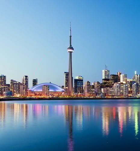 Toronto skyline at dusk with the CN Tower prominently lit. Skyscrapers surround it, reflecting vibrant lights on calm water, creating a serene urban scene.