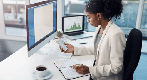 woman at a white modern desk with coffee and a computer looking at her mobile device