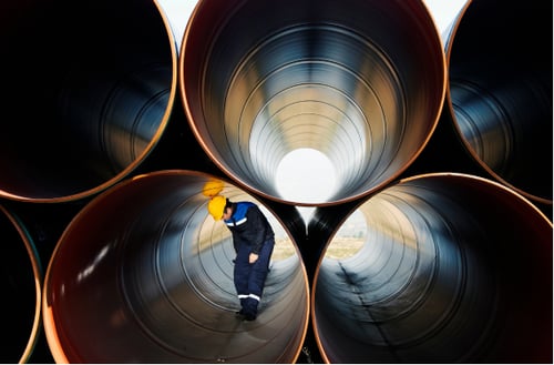 A worker in a blue uniform and yellow hard hat inspects large metal pipes stacked in a row. The scene conveys a sense of industrial focus and scale.