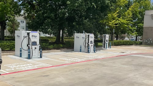 Several EVgo fast charging units installed in marked parking spaces at a public charging station, with EVgo logos on the chargers, protective bollards around each unit. Warehouse/large building in the right corner.