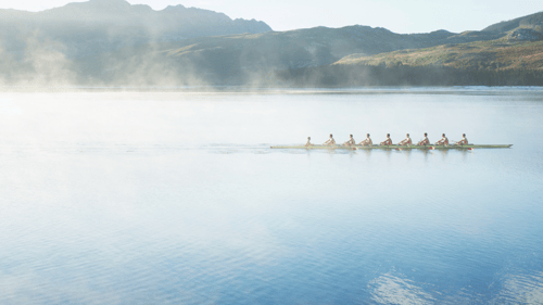 A rowing team of eight glides smoothly across a serene lake, surrounded by mist and distant hills. The image conveys tranquility and teamwork.