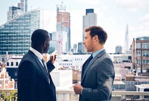 Two men in suits stand on a rooftop, discussing while looking at a city skyline with modern skyscrapers. The scene conveys a professional tone.