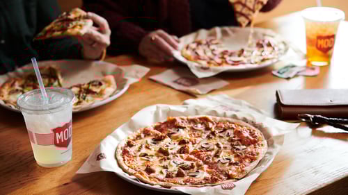 A table at a MOD Pizza restaurant with 3 different individual pizzas and some drinks. 2 people are eating their pizzas.