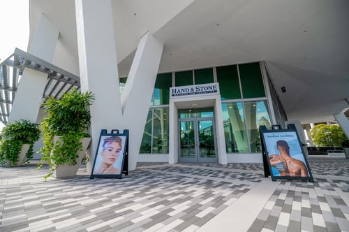 Outdoor plaza with patterned tile flooring in front of Hand & Stone spa entrance, showing portable massage setup and promotional signage