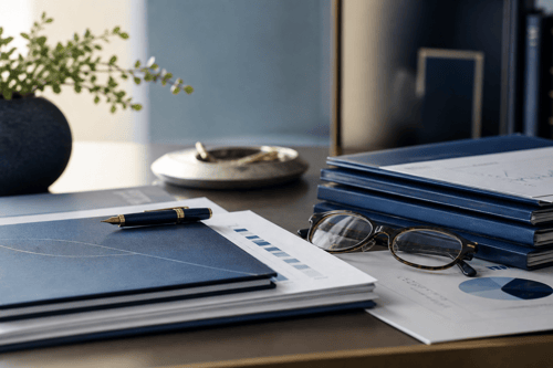 Navy blue presentation folders with financial charts and reports stacked on a polished desk beside reading glasses and a fountain pen.