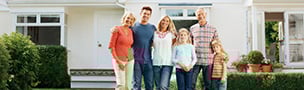 A family of six stands smiling in front of a white house with greenery. They look relaxed and happy, suggesting a warm and welcoming atmosphere.