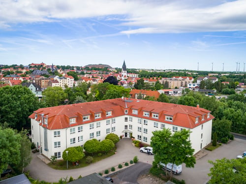 Luftaufnahme eines Seniorenwohnheims mit rotem Dach und heller Fassade, umgeben von Bäumen und einer Kleinstadt mit Kirche im Hintergrund. Mehrere Autos stehen vor dem Gebäude.