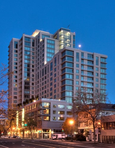 A modern multi-story building with glass windows is illuminated at dusk at The Grand Apartments, Oakland, CA
