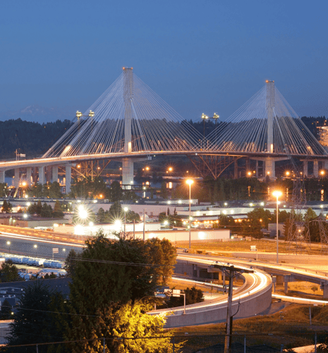 Elevated view of a suspension bridge at twilight, illuminated by streetlights. Curved roads and lush greenery create a serene, urban landscape.