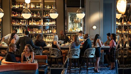 Elegant bar with guests socializing, bartenders preparing drinks, plush seating and shelves stocked with liquor bottles under warm lighting
