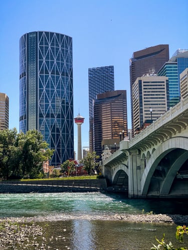 Skyline with modern skyscrapers, including one with a lattice design, a red observation tower, and a bridge. River in the foreground under a clear sky.