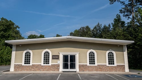 The Church of Jesus Christ of Latter-day Saints in Lafayette, Georgia. The viewer is presented with light-colored stucco and stone building with white trims. The front entryway is made of two class doors with two sets of windows in white trimming on either side. The name of the Church, "The Church of Jesus Christ of Latter-day Saints" is mounted to the left of the entryway.