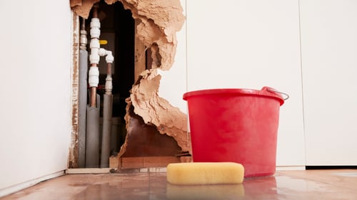 Interior view of a damaged white wall with a large hole exposing plumbing pipes, a red bucket catching drips, and water damage restoration in progress.
