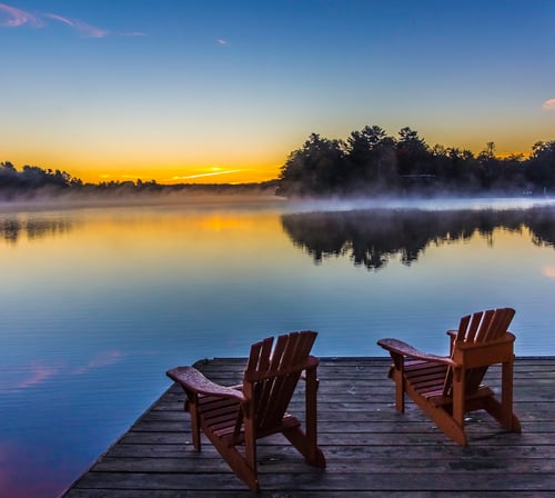 Two wooden chairs on a dock overlook a serene lake at sunrise, with mist rising from the water and a tranquil sky transitioning from orange to blue.