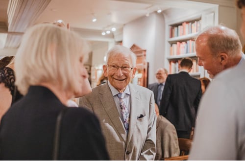 A smiling elderly man in a suit engages in conversation at a social gathering. People surround him in a warmly lit room with shelves of colorful books.