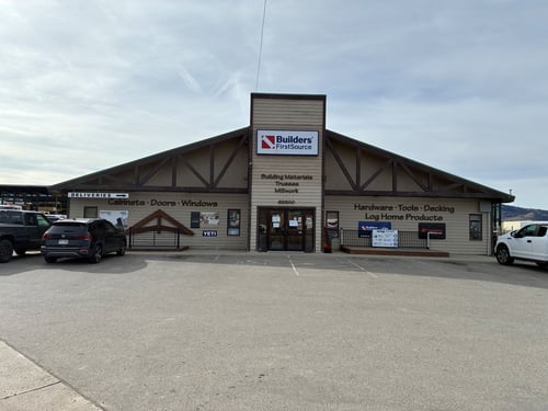 Front view of a Builders FirstSource store with a peaked roof and wood-trimmed exterior. The building has signage for cabinets, doors, windows, hardware, tools, decking, and log home products. Several vehicles are parked in front, and the main entrance displays the Builders FirstSource logo above the door.