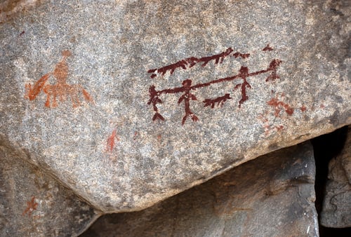 Ancient rock art depicting stick figures, possibly hunting, on a rocky surface. A figure rides an animal to the left; earthy tones dominate the scene.