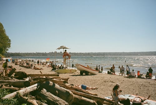 A sunny beach scene with people relaxing on the sand and swimming in the sea. A lifeguard sits on an elevated chair, overseeing the area. The mood is lively and relaxed. Logs are scattered on the sand in the foreground.