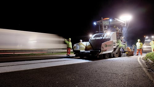 Road crew paving a highway at night using an asphalt paver under bright work lights.