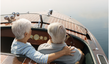 An elderly couple with gray hair sits closely on a wooden boat, facing a calm, misty lake. The scene conveys tranquility and companionship.