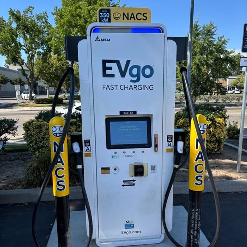 NACS EVgo fast charging unit installed in marked parking space at a public charging station, with EVgo logos on the charger,  and yellow protective bollards around the unit.