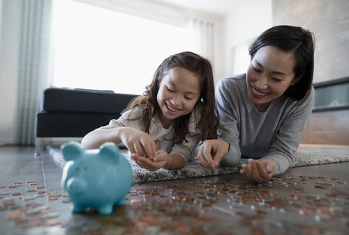 A woman and child lie on the floor, smiling as they count coins together. A blue piggy bank sits nearby, creating a warm, educational atmosphere.