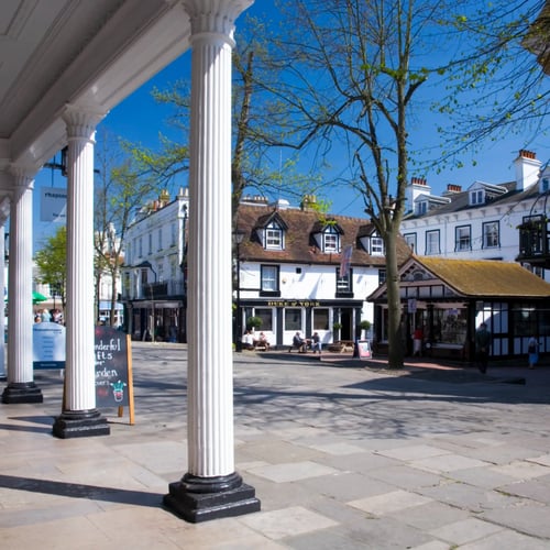 Knight Frank Tunbridge Wells Estate Agents - A sunny, pedestrianized historic square at The Pantiles in Royal Tunbridge Wells, featuring a white-pillared colonnade walkway, traditional tiled buildings, and trees.