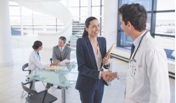 A doctor in a white coat shakes hands with a smiling businesswoman in an office. Two colleagues sit at a table in the background, suggesting a professional meeting.