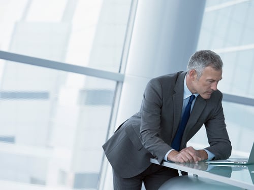 A man in a gray suit leans over a desk, focused on a laptop in a modern office with large windows. The scene conveys a professional and concentrated atmosphere.