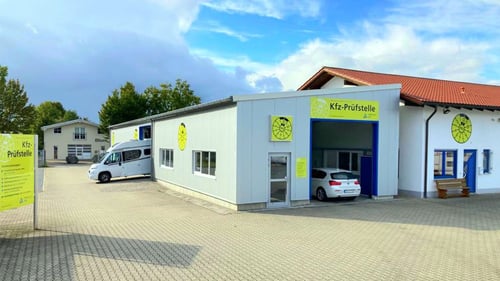 Außenansicht der Kfz‑Prüfstelle Steinach mit gelben Schildern, Hof und parkendem weißen Auto in der Prüfhallen-Einfahrt.