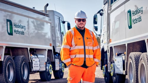 Worker wearing a hard hat and reflective orange safety clothing stands in front of Heidelberg Materials vehicles