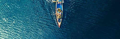 Aerial view of a small, colorful fishing boat with two people, surrounded by vast, rippling blue water. The scene conveys peacefulness and solitude.