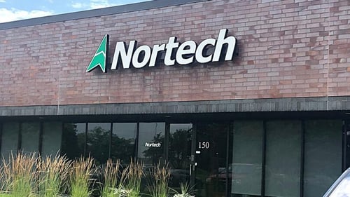 Front entry of building; floor to ceiling windows and doors stretch the length of the exterior wall.  Above the glass, brown brick extends to roof, displaying Nortech logo; green compass and white letters