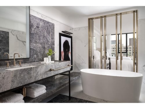 Luxurious modern bathroom with marble finishes, a freestanding bathtub, large mirror, and stylish decor, featuring a glimpse into a bedroom through sliding glass doors.