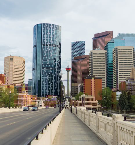 Urban skyline view of Calgary, featuring modern skyscrapers with varied architecture. The Calgary Tower stands prominently in the center. A bridge in the foreground leads into the bustling downtown area, under a partly cloudy sky.