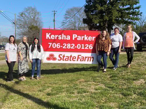 Six team members standing and smiling together wearing professional attire next to red and white State Farm sign