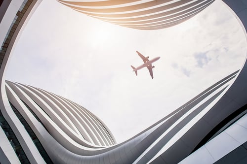A plane flies above a futuristic building with curved, wavy lines and a bright sky. The perspective creates a sense of openness and modernity.