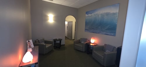 Spa waiting area with comfortable chairs, large ocean wave art, warm lighting, and salt lamp at Hand & Stone.