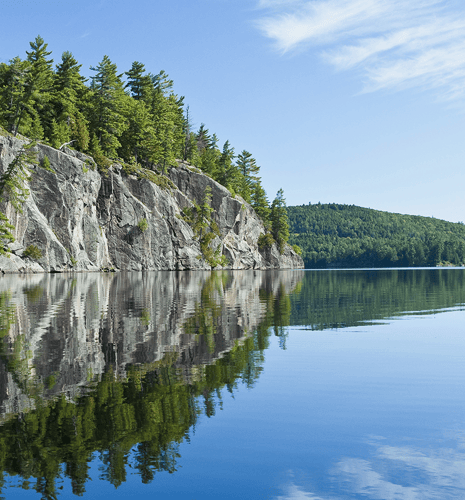 A tranquil lake scene featuring rocky cliffs and lush trees reflected in calm waters under a clear blue sky.