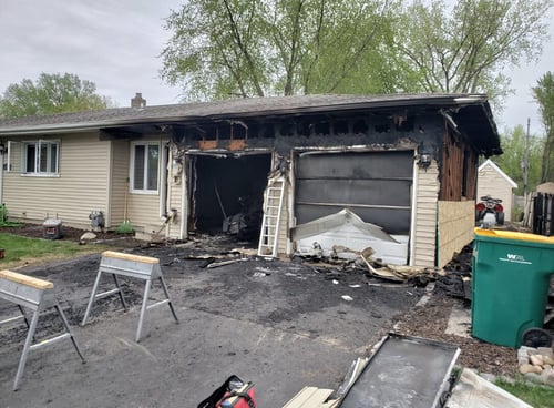 A partially charred home stands surrounded by restoration equipment, with construction materials and workers on site as the property undergoes repairs to recover from fire damage