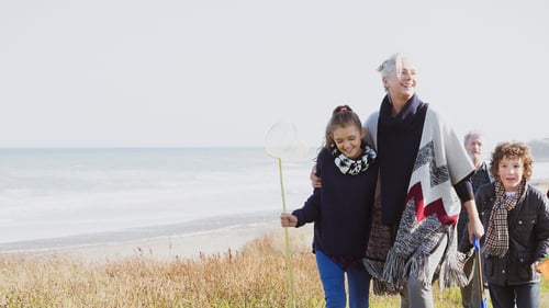 A joyful family strolls along a grassy beach path, with the ocean in the background. A young girl holds a fishing net, smiling alongside an older woman.