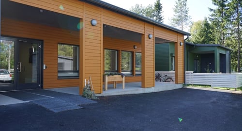 Kuvassa on moderni yksikerroksinen rakennus, jossa on puinen julkisivu maalattu oranssilla ja vihreällä. Rakennuksessa on suuret ikkunat, tasakatto ja esteetön sisäänkäynti ilman näkyviä portaita. Lisäksi on pieni kuisti alue penkkeineen. Ympäröivä alue vaikuttaa hyvin hoidetulta päällystetyillä pinnoilla.