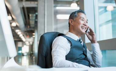 A man in a suit smiles while talking on the phone, seated at a desk in a modern, bright office with large windows and industrial-style lighting.