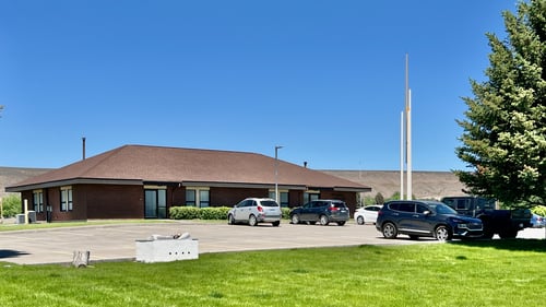 The outside of a church building. There is a separate steeple on the corner of the building. A parking lot with automobiles, grass, and a large pine tree surround the church.