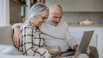 An elderly couple sits close on a couch, smiling while using a laptop. The cozy setting conveys warmth and connection, with soft, neutral tones.