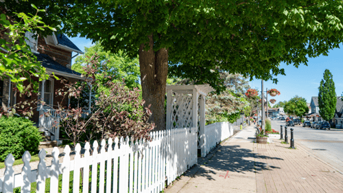A quaint suburban street in Vaughan, Ontario features a traditional white picket fence, lush green trees, vibrant hanging flower baskets, and cozy homes under a clear blue sky.