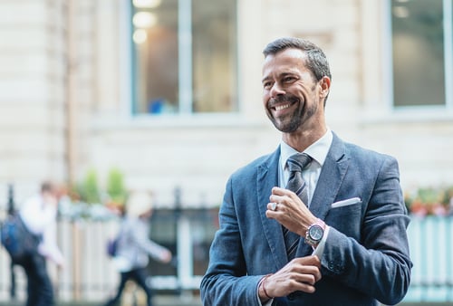 Smiling man in a suit adjusts his sleeve outdoors, exuding confidence. Blurred cityscape and pedestrians in the background convey busy urban life.