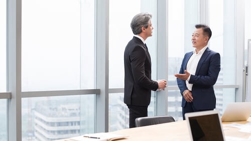 Two businessmen in suits engage in conversation in a bright office with large windows overlooking a cityscape. A laptop rests on a wooden table.